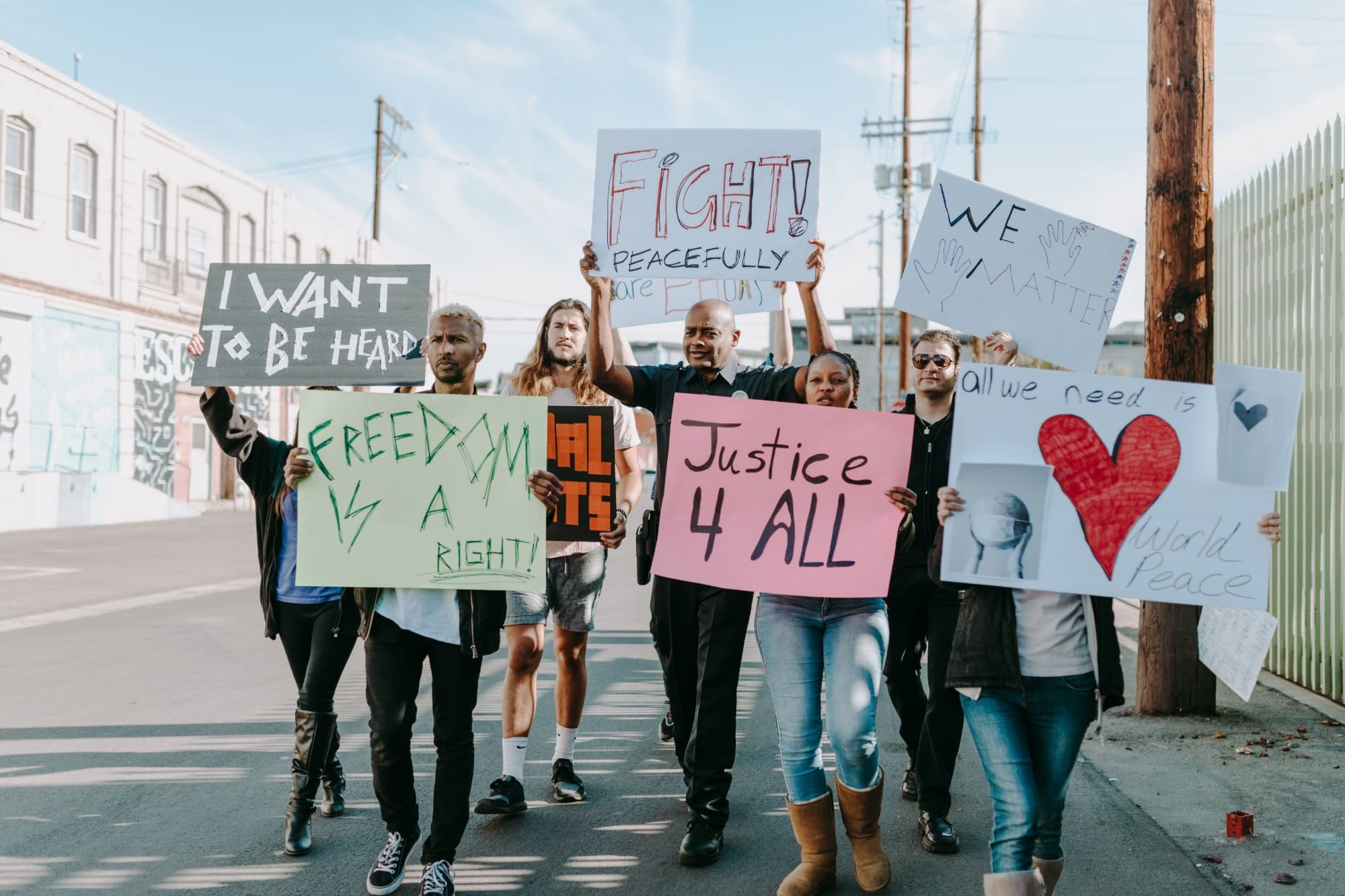 People marching down a sunny street holding hand-made signs: Justice 4 All, Freedom is a right, We matter